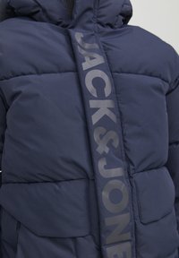 Merihenkinen puhvijakku, jossa on kiiltävä "JACK & JONES" -logonauha, tikattu pinta ja huppu. Mukana etupocketit säilytystä varten.