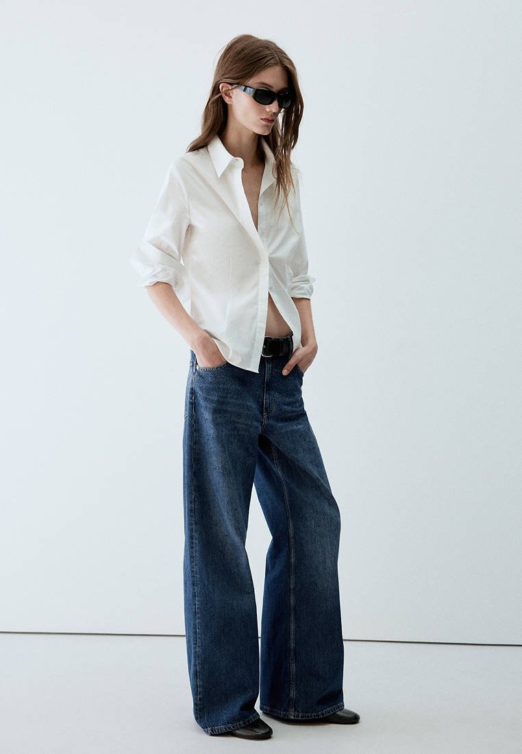 Femme portant des lunettes de soleil noires, une chemise blanche boutonnée aux manches retroussées, un jean bleu à larges jambes et des chaussures noires, debout les mains dans les poches.