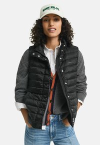 Schwarze Puffer-Weste über einem grauen Strickpullover, kombiniert mit einer mehrfarbigen, geknöpften Strickjacke. Eine cremefarbene Kappe mit grüner Schrift rundet den Look ab.