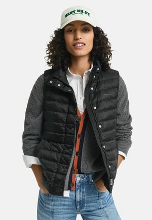 Black puffer vest over a gray knit sweater, paired with a multicolored buttoned cardigan. A cream cap with green text completes the look.