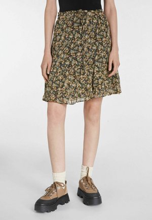 Femme portant une jupe noire à fleurs mi-longue avec de petites fleurs, des chaussures de randonnée beiges à semelles épaisses, et des chaussettes blanches montant jusqu'à la cheville.