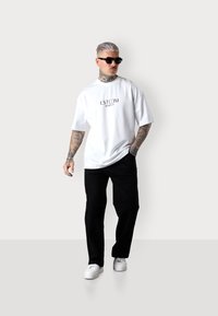 T-shirt en coton blanc avec le texte noir "ESTEEM MARBELLA", porté avec un pantalon noir et des baskets blanches. Le modèle a les cheveux courts argentés et des lunettes de soleil.