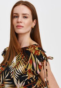 Schwarze Bluse mit buntem tropischem Blätterdruck, gerafften Ärmeln mit Bindebändern, leichtem Stoff und Akzenten in Rot, Grün und Pink.