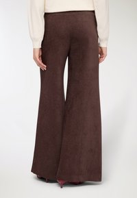 Pantaloni marroni a zampa d'elefante in tessuto morbido e strutturato con vita alta. Presentano un design a gamba larga che arriva fino a terra. Tacchi alti rossi parzialmente visibili.