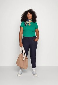 Mulher com cabelo encaracolado a usar uma blusa verde, calças azul-marinho, um lenço estampado, ténis brancos, e a segurar uma mala de pano bege, contra um fundo branco.