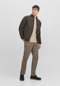 Brun quiltad jacka med ståkrage, kombinerad med en beige tröja och beige byxor. Modellen bär svarta skor med texturerad yta.