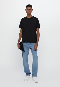 Camiseta de algodón negra con logo de la marca en relieve, combinada con jeans de mezclilla azul claro y zapatillas blancas, sosteniendo un accesorio negro.