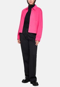 Rosa Cropped-Sweatshirt mit spitzem Kragen, Frontknöpfen und Seitentaschen, kombiniert mit schwarzen Jogginghosen mit Kordelzug und seitlichen Streifen.