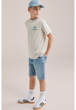 Chlapec v svetlo béžovom tričku s nápisom "Poďme objavovať", rifľových šortkách, pruhovaných bielych ponožkách, bielych teniskách a svetlomodrej šiltovke, ktorý hľadí do strany.