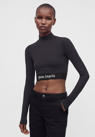 Γυναίκα με ξυρισμένο κεφάλι, φοράει μαύρο crop top με μακριά μανίκια και το κείμενο "palm angels" και μαύρο παντελόνι, στέκεται με το ένα χέρι στην τσέπη.