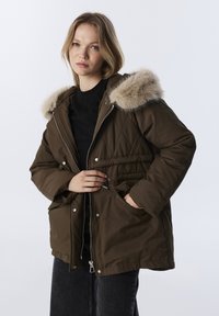 Bruine, gewatteerde parka met een nepbont gevoerde capuchon, rits- en drukknoopsluiting, verstelbare taille en zijzakken. Zwarte top en donkere jeans eronder.