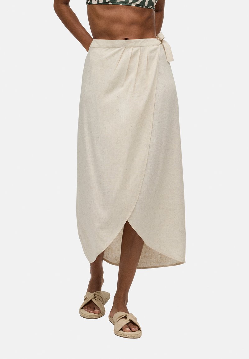 Falda envolvente de lino beige con detalle de lazo en la cintura, con un dobladillo asimétrico y un corte relajado. Combinada con sandalias informales de tipo slip-on.