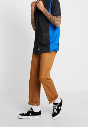 Mann trägt schwarzes und blaues kurzärmeliges Hemd, braune hochgekrempelte Hose, weiße Socken und schwarze Turnschuhe mit weißem Stern, steht vor weißem Hintergrund.