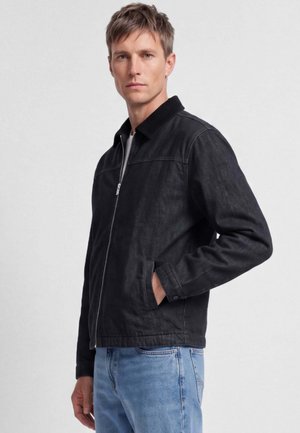 Mann trägt eine schwarze Reißverschlussjacke mit Kragen und hellblaue Jeans, steht mit einer Hand in der Tasche vor einem schlichten Hintergrund.
