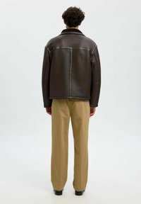 Brun läderjacka med klassisk krage, med en slät текстur och vidskuren design, ihopparad med beiga raka byxor.