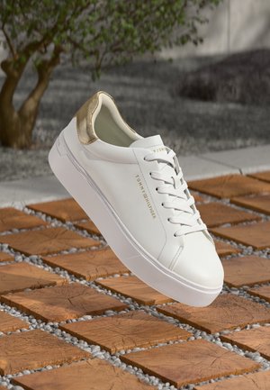 Biała tenisówka Tommy Hilfiger ze złotym detalem na pięcie, białe sznurówki, wyświetlona pod kątem na drewnianych kwadratowych płytkach z małymi szarymi kamykami.