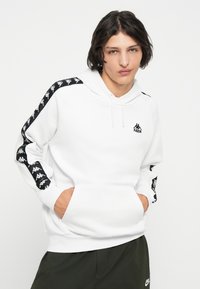 Sudadera blanca con logo negro en el pecho y franjas laterales negras con patrón. Mangas largas con puños de canalé. Bolsillo tipo canguro en la parte delantera.