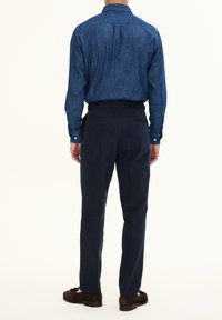 Denim button-up shirt in dark blue, paired with tailored navy trousers and brown suede shoes, viewed from the back.