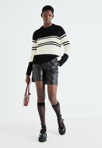 Maglione a maglia cable nera e crema con tre righe orizzontali, abbinato a pantaloni corti in pelle nera e scarpe con plateau spesse.