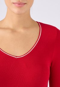Maglietta rossa a maglia con scollatura a V profonda, caratterizzata da un bordo bianco a contrasto lungo il colletto. Tessuto liscio, design aderente.