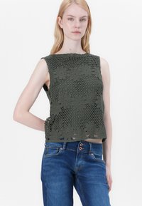 Oliven grønstrikket topp med blomsterdesign, uten ermer og løs passform, kombinert med blå jeans som har knappdetaljer.