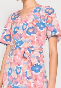 Blumenkleid in Pastellfarben mit V-Ausschnitt, kurzen Ärmeln und gebundenem Taillenbereich. Der Stoff zeigt große blaue und rosa Blumenmuster.