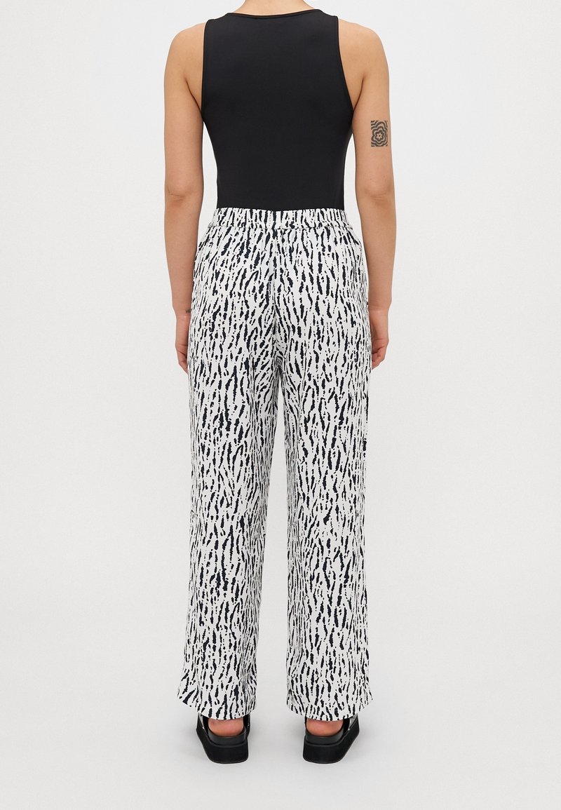 Femme portant un haut noir sans manches et un pantalon blanc à larges jambes avec un motif abstrait noir, debout devant un fond blanc uni.