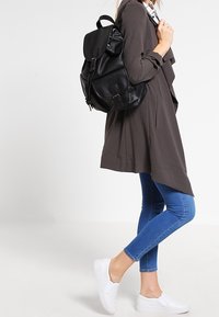 Czarny skórzany plecak, szary kardigan w warstwach, niebieskie skinny jeans, białe buty na slip-on. Gładkie tekstury; minimalistyczny design.