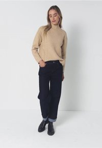 Beige gestrickter Rollkragenpullover mit strukturiertem Muster, kombiniert mit dunkelblauen Jeans und schwarzen Stiefeletten mit umgeschlagenen Säumen. Einfaches Design, lockere Passform.