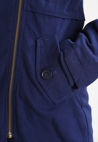 Giacca blu navy con chiusura a zip e dettaglio della tasca con bottone. Realizzata in tessuto testurizzato, presenta una zip e cuciture in oro a contrasto.
