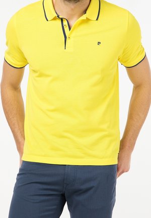 Polo jaune avec une patte à deux boutons, bordure bleu marine sur le col et les manches, et un petit logo sur la poitrine gauche. Porté avec un pantalon bleu.