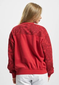 Roter Pullover mit einem strukturierten Blumenmuster auf dem oberen Rücken und den Ärmeln. Glattes Material, gerippter Bund und Ärmelbündchen.