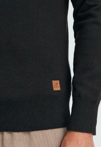 Pull en tricot gris foncé avec des poignets et un ourlet côtelés, présentant une petite étiquette en cuir marron près du bas avec des détails de logo.
