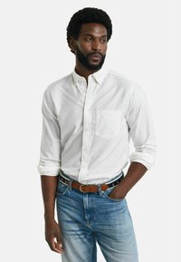 Ein weißes Langarmhemd mit Knopfleiste verfügt über eine Brusttasche und ist mit hellblauen Jeans sowie einem braunen Gürtel mit einem gemusterten Akzent kombiniert.