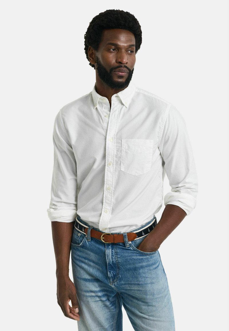 Ein weißes Langarmhemd mit Knopfleiste verfügt über eine Brusttasche und ist mit hellblauen Jeans sowie einem braunen Gürtel mit einem gemusterten Akzent kombiniert.
