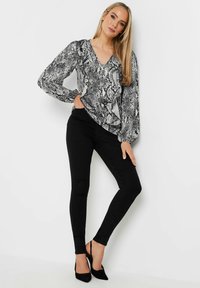 Blusa nera con stampa serpente, dettagli arricciati, scollatura a V e maniche voluminose, abbinata a jeans skinny neri e tacchi neri.