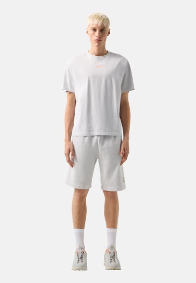 Jeune personne aux cheveux blonds courts portant un t-shirt blanc ample, un short blanc assorti, des chaussettes blanches et des baskets blanches, debout face à l'avant.
