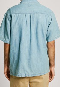 Camisa de manga corta azul claro en tejido suave, con un cuello estándar, pliegue en la parte posterior y dobladillo recto, con detalles de costura visibles.