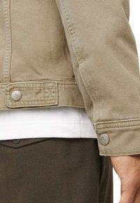 Veste en denim beige avec un poignet boutonné, une poche latérale et des coutures décoratives. Portée par-dessus une chemise blanche et un pantalon sombre.