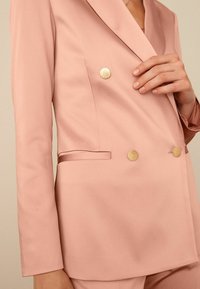 Vaaleanpunainen kaksirivinen bleiseri kultaisilla napeilla, satiinimainen tekstuuri, tyylikkäät kaulukset ja sivusomisteet, suunniteltu modernia istuvuutta varten.