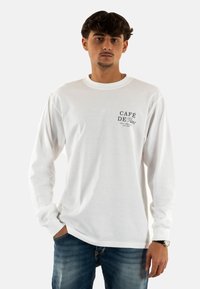 Camiseta blanca de manga larga hecha de algodón, con un logo impreso en negro "Café de Paris" en la parte superior izquierda, combinada con unos vaqueros azules.