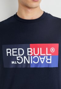 Tamsiai mėlynas medvilninis marškinėliai su grafiniu dizainu, kuriame balta spalva parašyta "RED BULL", raudonai "RACING" ir tamsiai mėlynas fonas.