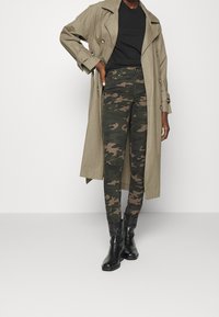 Beige trenchcoat, svart t-shirt och kamouflagemönstrade skinny jeans. Svarta ankelstövlar. Fokus på tygtexturer och mönster.