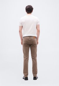 Άνδρας με την πλάτη, φοράει λευκό μπλουζάκι, καφέ τζιν και μαύρα παπούτσια, στέκεται μπροστά σε απλό ανοιχτό φόντο.