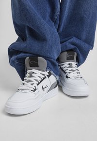 Baskets blanches avec des accents gris, une languette texturée et un logo noir. Associées à un pantalon en denim bleu large, offrant une coupe décontractée et un ourlet effiloché.