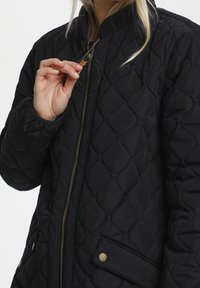 Chaqueta de cuirette negra con cierre de cremallera en la parte delantera, cuello acanalado, dos bolsillos laterales y acentos en hardware tono dorado. Textura suave y acolchada.