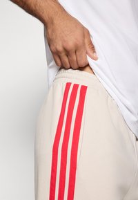 Bežinės sportinės kelnės su raudonomis vertikaliomis juostelėmis šonuose, turinčios elastingą juosmenį ir minkštą, tekstūruotą audinį.