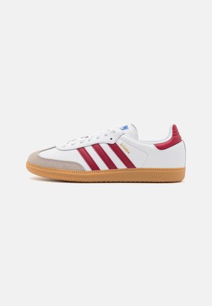 Zapatilla blanca con rayas rojas, suela de goma y puntera de ante gris claro. Presenta una etiqueta azul, cordones blancos y una marca en el lateral.