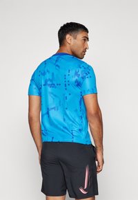 Camiseta atlética azul con un patrón texturizado y diversas tonalidades de azul. Combinada con pantalones cortos negros que muestran un logo rosa.
