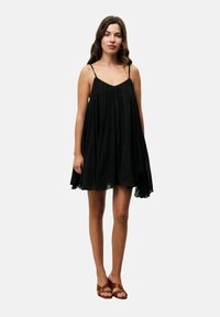 Zwarte mouwloze jurk met een vloeiende silhouette, gathering bij de buste en een zachte textuur. Draagt met bruine sandalen met banden.